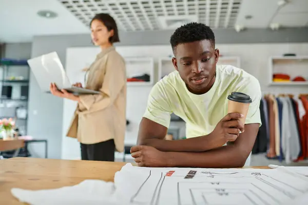 stock image black man with paper cup looking at clothes with format sizes near asian designer with laptop