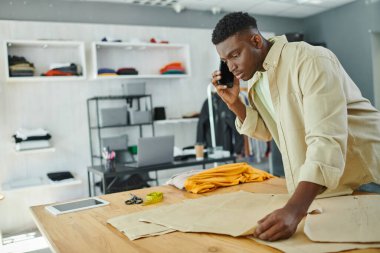 Ciddi bir Afro-Amerikan tasarımcı dikiş modellerine bakıyor ve atölyedeki akıllı telefondan konuşuyor.