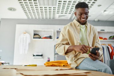 gülümseyen Afro-Amerikan tasarımcı akıllı telefondan baskı stüdyosundaki dikiş desenlerinin yanında mesaj atıyor.