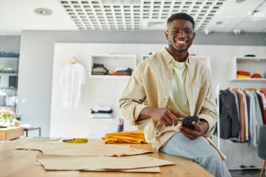 Neşeli Afrikalı Amerikalı tasarımcı cep telefonuyla baskı stüdyosunda dikiş desenlerinin yanında oturuyor.