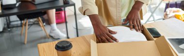 Afro-Amerikan bir adamın atölyedeki kağıt bardağın yanında kıyafet paketlediği görüntüler.