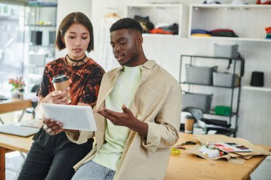 ambitious interracial designers with digital tablet and paper cup working on startup in print studio clipart