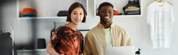 happy asian woman and african american man with laptop smiling at camera in print studio, banner