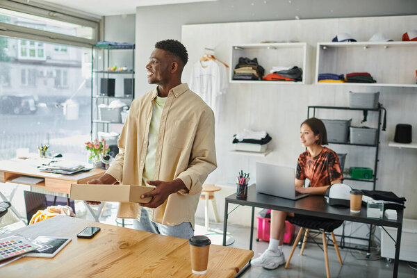 довольный африканский американец с посылкой рядом с устройствами и азиатский дизайнер за ноутбуком в типографии