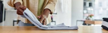 cropped view of african american worker of textile print studio folding clothing neatly, banner clipart