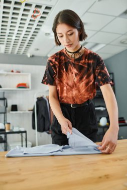 young and trendy asian woman folding clothes on table in textile print studio, small business clipart