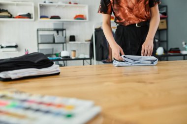 partial view of experienced woman folding clothes on table in textile print atelier, small business clipart