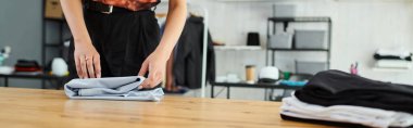 cropped view of young woman folding clothes neatly on table in textile print atelier, banner clipart