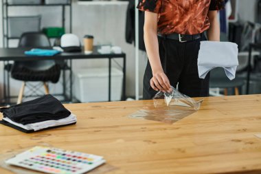 cropped view of young woman packing folded clothes in plastic bag on table in print workshop clipart