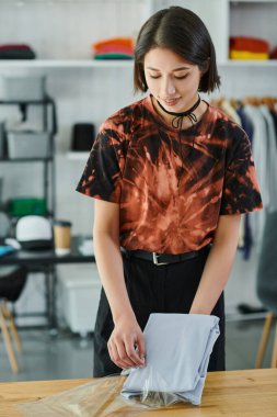 Gülümseyen Asyalı kadın özenle katlanmış kıyafetlerini naylon poşete koyup baskı stüdyosunda moda tasarımı yapıyor.