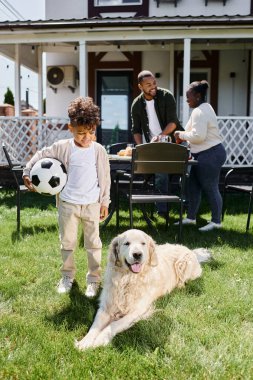 Aile zamanı, mutlu Afro-Amerikan çocuk arka bahçede ailesinin ve köpeğinin yanında futbol oynuyor.