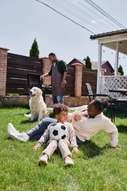 Mutlu Afro-Amerikan annesi köpeği yanında oğluyla çimenlerde otururken kocası da Bbbq Grill 'de yemek yapıyor.