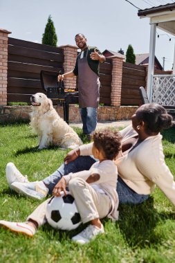 african american mother sitting on lawn with son near dog while happy husband cooking on bbq grill clipart