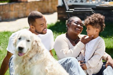 Afro-Amerikan ailesinin gülümseyen ve köpek yakınındaki çimlerde oturan aile portresi.