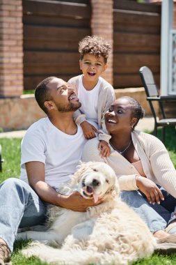 quality time of happy african american parents and son smiling and sitting on grass near dog clipart