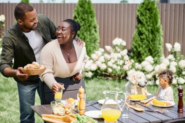 happy african american married couple serving table in garden near son playing with dog on backyard clipart