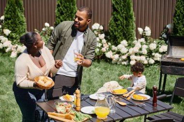 happy african american parents serving table in garden near son playing with dog on backyard clipart