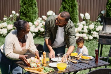 Neşeli Afrikalı Amerikalı ebeveynler bahçede masa servisi yapıyor. Oğulları arka bahçede oynuyor, aileleri de.