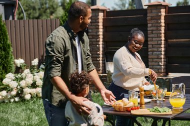 Mutlu Afro-Amerikan ebeveynler bahçede masa servisi yapıyorlar. Oğullarının bahçesinde futbol topu tutuyorlar.
