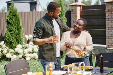 Afrikalı Amerikalı adam mutlu karısına sarılıyor cam kasede salata karıştırıyor arka bahçede de bbq yiyor.