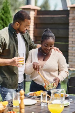 Mutlu Afro-Amerikalı adam arka bahçede bbq yerken cam kasede salata tutarak karısına sarılıyor.