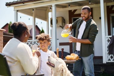 Neşeli Afrikalı Amerikalı çocuk arka bahçede aile bbq sırasında gülümsüyor ve köpek ve ebeveynlerin yanında duruyor.