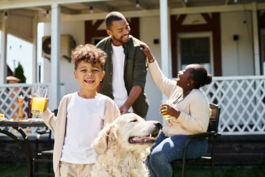Mutlu Afrikalı Amerikalı çocuk gülümseyip köpeğini okşarken ailesinin yanında bir bardak portakal suyu tutuyor.