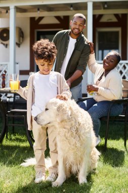 Neşeli Afrikalı Amerikalı çocuk gülümsüyor ve elinde bir bardak portakal suyu tutarken köpeğini okşuyor.