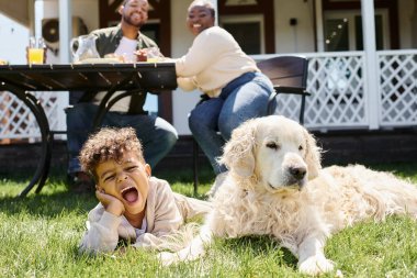 Duygusal Afrikalı Amerikalı çocuk yeşil çimlerde uzanmış köpek ve ailesinin bahçesinde öğle yemeği yiyor.