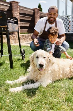 Önlüklü, gülümseyen Afro-Amerikan adam elinde yemek maşası ve oğlunun yanındaki köpeğe bakıyor.