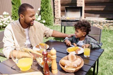 cheerful african american father eating lunch with curly son outdoors, grilled sausages and corn clipart