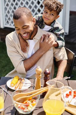 smiling african american child hugging excited father in braces on backyard of house, family time clipart