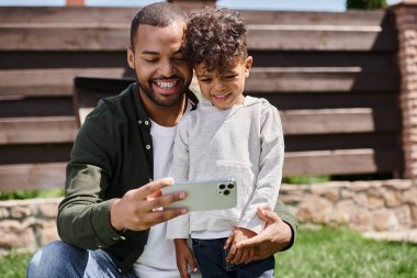 pleased african american father in braces taking selfie on smartphone with his son on backyard clipart