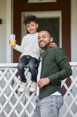 Diş teli takmış, elinde portakal suyu tutan ve verandada oturan oğlunu kucaklayan mutlu Afro-Amerikan babası.