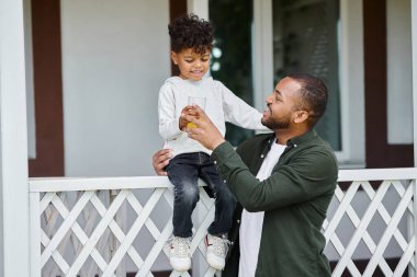 happy african american father in braces holding orange juice and hugging cute son sitting on porch clipart