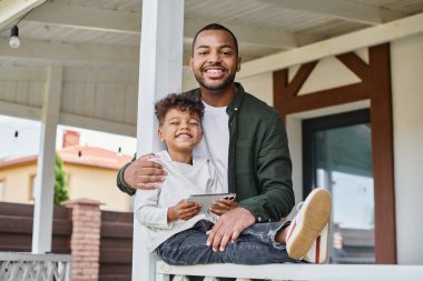Neşeli Afrikalı Amerikalı baba oğluna sarılıyor verandada oturuyor ve elinde akıllı telefon, siyah aile