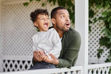 astonished african american father and son looking away surprisingly while sitting near white fence clipart
