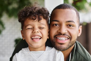 Mutlu Afro-Amerikan bir adamın aile portresi. Diş teli takmış kameraya bakarken kıvırcık oğluna sarılıyor.