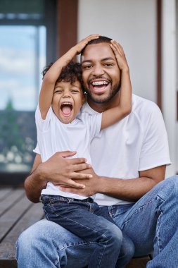 Heyecanlı Afrikalı Amerikalı çocuk modern evin verandasında otururken mutlu babaya sarılıyor.