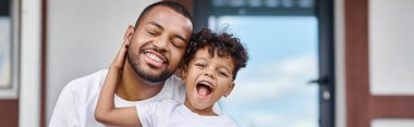 excited african american boy hugging jolly father in braces while sitting on porch of house, banner clipart