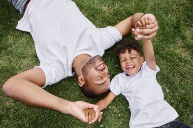 top view of happy african american father and son lying on lawn on backyard and holding hands clipart