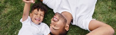 top view of positive african american father and son lying on green lawn on backyard, family banner clipart