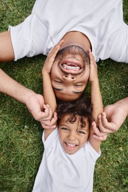 top view of african american father and son lying on green lawn on backyard, happy family memories clipart