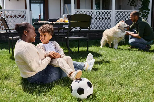 幸せなアフリカ系アメリカ人の母親は 裏庭のサッカー 犬の隣に息子と芝生に座っています — ストック写真