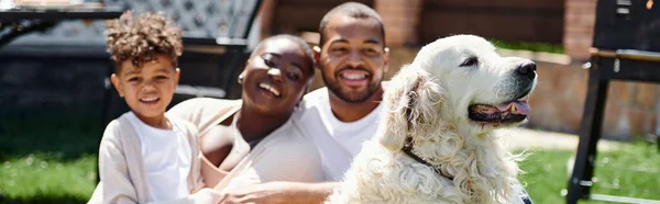 stock image family banner of joyful african american parents and son smiling and sitting on lawn near dog