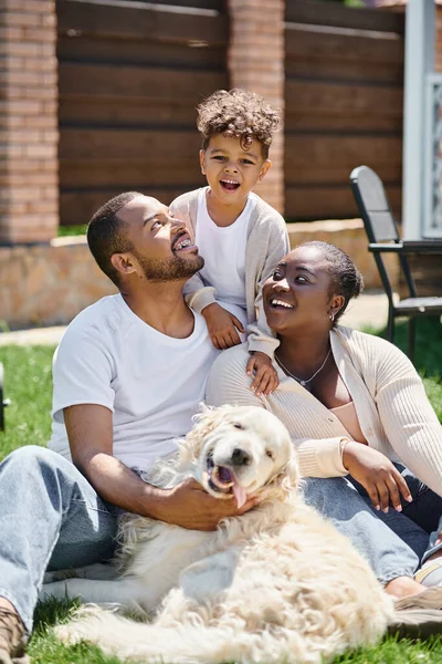幸せなアフリカ系アメリカ人の両親と息子の質の高い時間 笑顔と犬の近くの草の上に座って — ストック写真