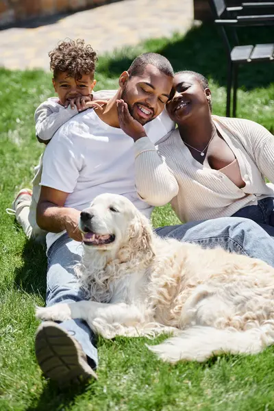 幸せなアフリカ系アメリカ人の両親と息子の肖像画 笑顔で犬の近くの緑の芝生に座って — ストック写真