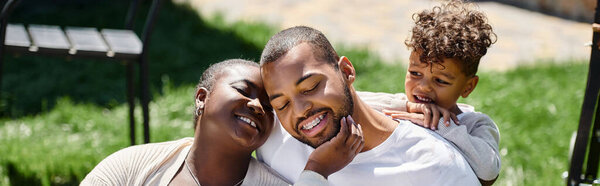 family moment of happy african american parents and son smiling and sitting on green lawn, banner