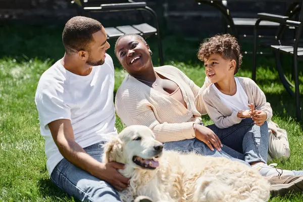 ジョリー アフリカ系アメリカ人の両親と息子が笑顔で犬の近くの緑の芝生に座る家族の瞬間 — ストック写真