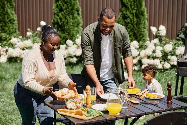 веселые африканские американские родители, обслуживающие стол в саду рядом сын играет на заднем дворе, общая еда